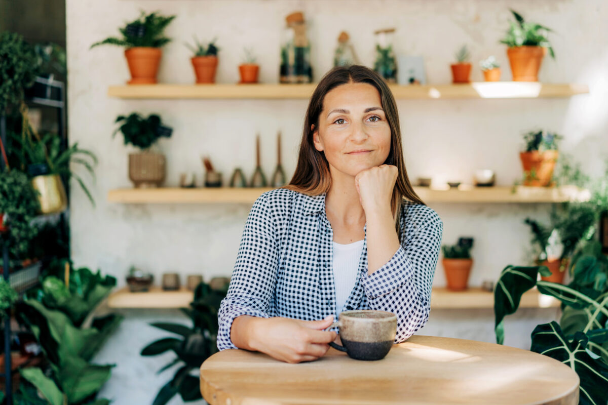 A woman sitting at a wooden table, holding a cup, surrounded by various potted plants on shelves in a bright, inviting space.
